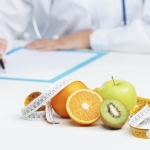 "Doktorun önünde masanın üstünde ölçüm bandı ile çevrili portakal, elma ve kivi bulunan bir görsel. Sağlıklı yemek yemek önemi ve ölçümün diyet üzerindeki rolünü temsil eder